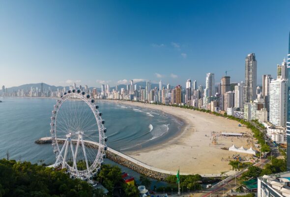 Vista panorâmica de Balneário Camboriú, com sua icônica roda-gigante à esquerda, próxima ao mar. A praia se estende ao longo da cidade, que é cercada por altos edifícios modernos. O céu está limpo, com poucas nuvens, e o mar está tranquilo.