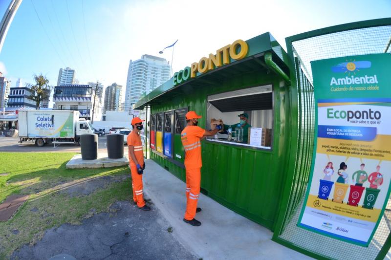 Colaboradores da Ambiental em uniformes laranja no Ecoponto de Itajaí, realizando coleta seletiva. Ao fundo, caminhão de coleta seletiva e prédios urbanos.