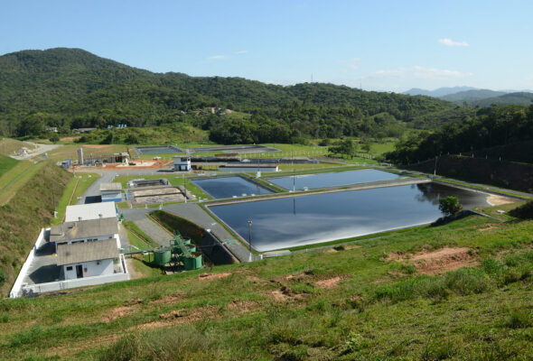 A imagem mostra uma estação de tratamento de líquidos percolados localizada em uma área cercada por colinas verdes e vegetação densa. A instalação inclui tanques de armazenamento, prédios técnicos e uma estrada pavimentada, tudo sob um céu azul claro.