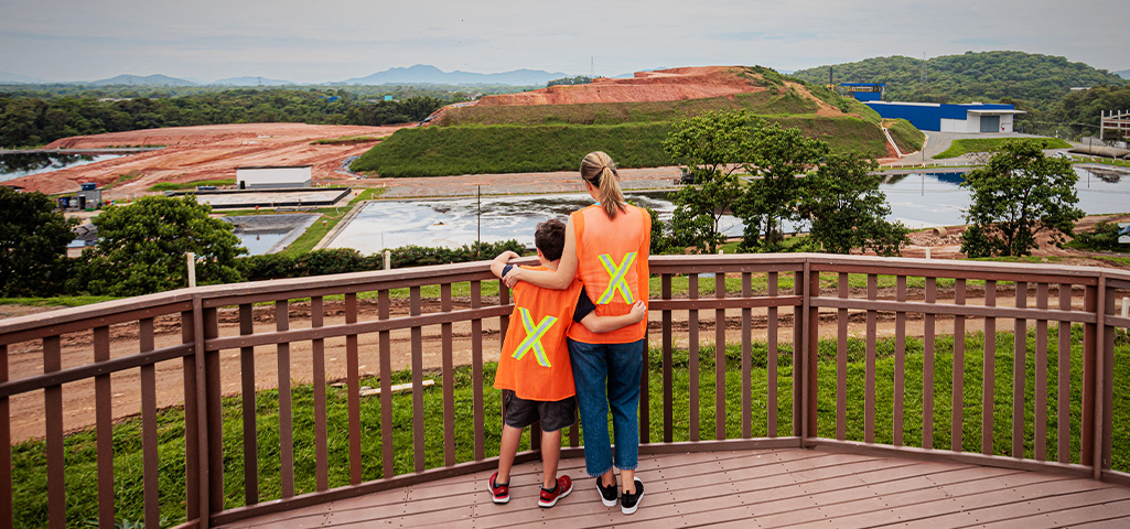 Mãe e filho em visita a uma usina de mineração de carvão, observando o horizonte de uma área industrial com caminhões e equipamentos de mineração, sob céu nublado.