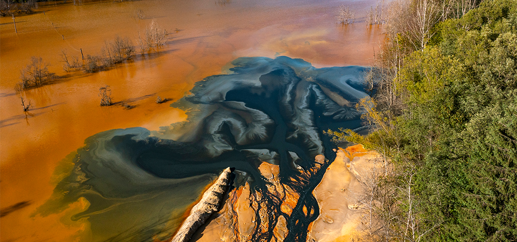 Imagem aérea de uma lagoa de cor laranja com áreas de água escura, rodeada por árvores verdes, destacando uma paisagem natural única e vibrante.