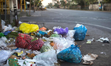 Lixo acumulado na calçada de uma rua, com sacolas plásticas de diferentes cores e resíduos espalhados, mostrando problema de descarte inadequado de lixo.
