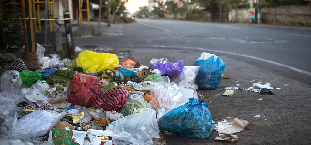Lixo acumulado na calçada de uma rua, com sacolas plásticas de diferentes cores e resíduos espalhados, mostrando problema de descarte inadequado de lixo.