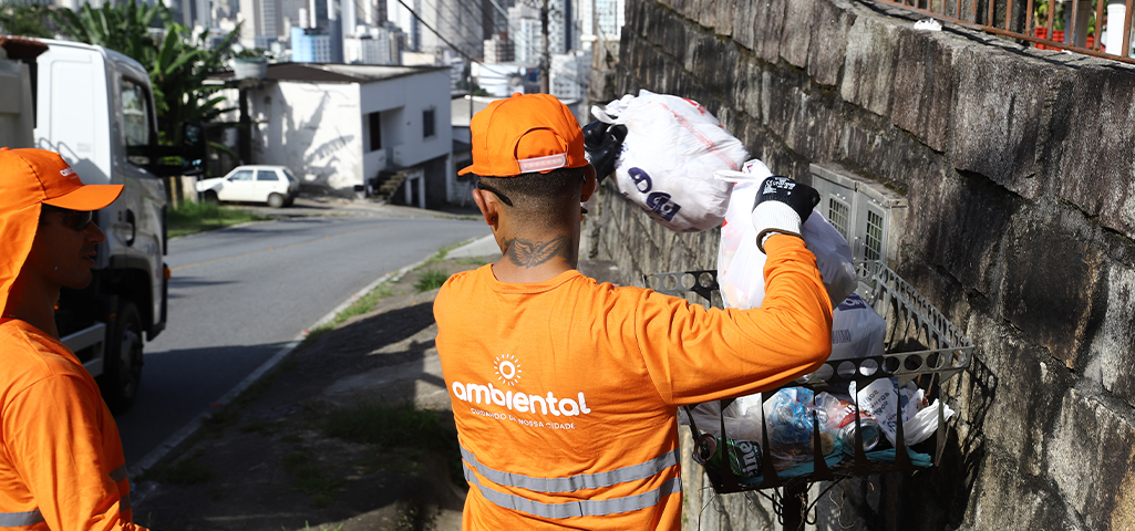 Funcionários de coleta de lixo atuando na limpeza urbana, coletando resíduos em uma área residencial com roupas laranja e equipamentos de proteção.